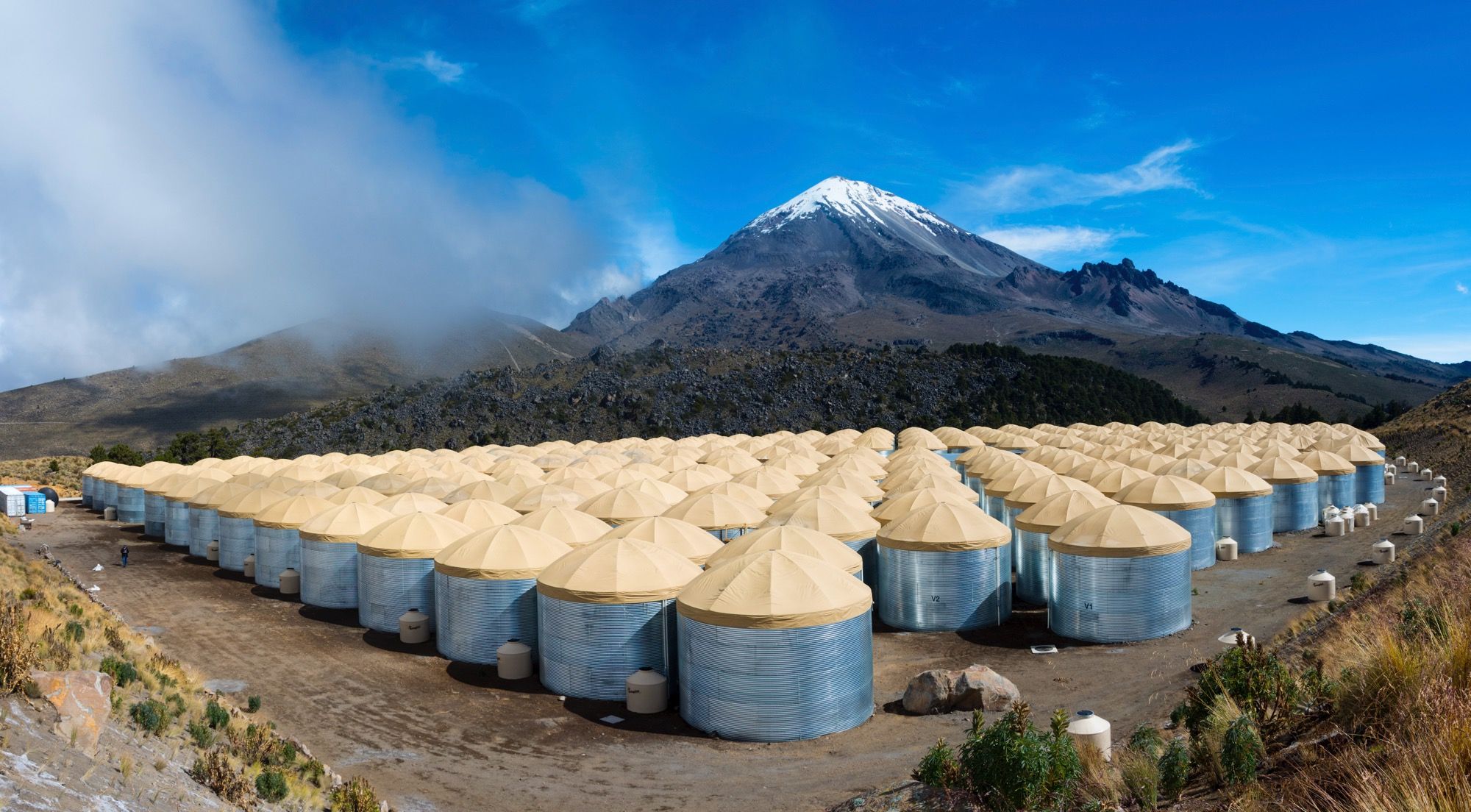 The water tanks of the HAWC observatory, steel cylinders with canvas roofs laid out in a compact grid on the slope of the volcano, with mountains in the background.